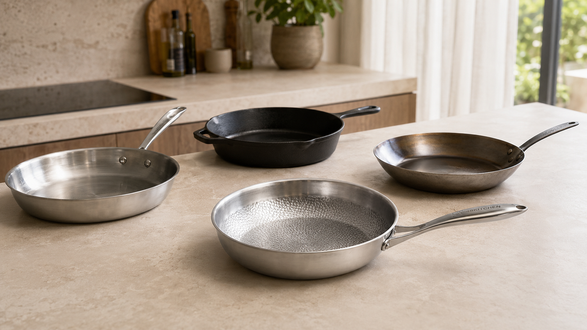> Verschillende koekenpanmaterialen naast elkaar, met een titanium-versterkte pan, RVS, gietijzer en koolstofstaal.